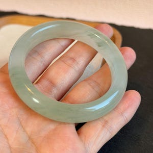 K&ouml;nnte beinhalten: Ein hellgr&uuml;nes Jade-Armreif-Armband. Die glatte, polierte Oberfl&auml;che reflektiert das Licht und zeigt die nat&uuml;rlichen Farbvariationen. Das Armband wird in einer offenen Hand gehalten, wodurch seine kreisf&ouml;rmige Form und sein elegantes Design hervorgehoben werden.