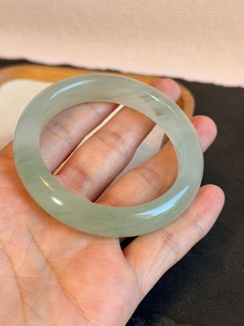 K&ouml;nnte beinhalten: Ein hellgr&uuml;nes Jade-Armreif-Armband. Die glatte, polierte Oberfl&auml;che reflektiert das Licht und zeigt die nat&uuml;rlichen Farbvariationen. Das Armband wird in einer offenen Hand gehalten, wodurch seine kreisf&ouml;rmige Form und sein elegantes Design hervorgehoben werden.