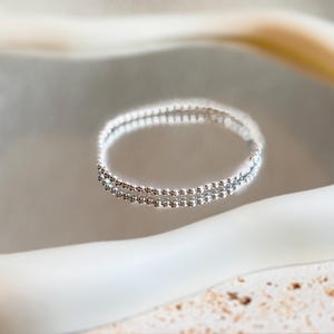 May include: A delicate silver bracelet with two rows of small, sparkling beads. The bracelet is displayed on a reflective surface, creating a mirrored effect. The background is a soft, neutral color, highlighting the jewelry.