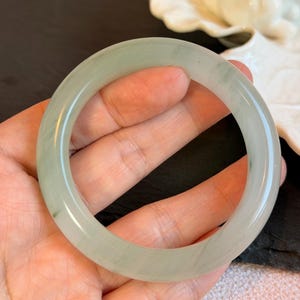 K&ouml;nnte beinhalten: Ein hellgr&uuml;nes Jade-Armreif-Armband. Das Armband hat eine runde Form mit einer glatten, polierten Oberfl&auml;che. Die Farbe ist ein weiches Gr&uuml;n mit nat&uuml;rlichen Variationen. Das Armband ist eine Standardgr&ouml;&szlig;e und scheint aus einem durchscheinenden Stein zu bestehen.