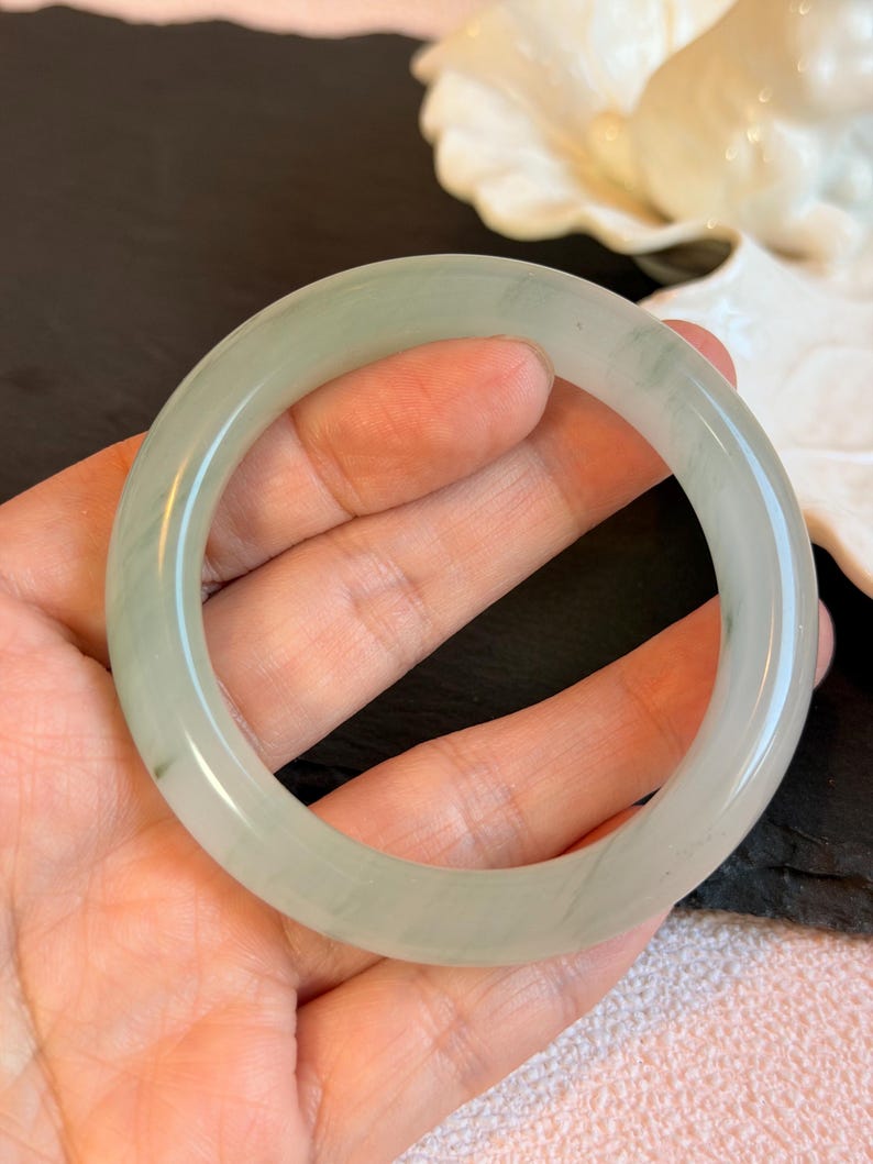 K&ouml;nnte beinhalten: Ein hellgr&uuml;nes Jade-Armreif-Armband. Das Armband hat eine runde Form mit einer glatten, polierten Oberfl&auml;che. Die Farbe ist ein weiches Gr&uuml;n mit nat&uuml;rlichen Variationen. Das Armband ist eine Standardgr&ouml;&szlig;e und scheint aus einem durchscheinenden Stein zu bestehen.