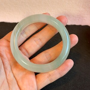 K&ouml;nnte beinhalten: Ein durchscheinendes, hellgr&uuml;nes Jade-Armreif-Armband. Die glatte, polierte Oberfl&auml;che reflektiert das Licht und zeigt die nat&uuml;rlichen Variationen des Steins. Das kreisf&ouml;rmige Design ist ein klassischer Schmuckstil.