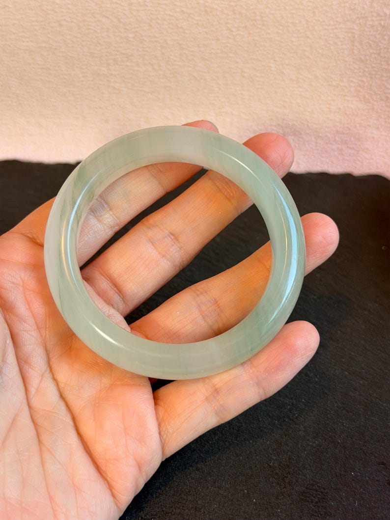 K&ouml;nnte beinhalten: Ein durchscheinendes, hellgr&uuml;nes Jade-Armreif-Armband. Die glatte, polierte Oberfl&auml;che reflektiert das Licht und zeigt die nat&uuml;rlichen Variationen des Steins. Das kreisf&ouml;rmige Design ist ein klassischer Schmuckstil.