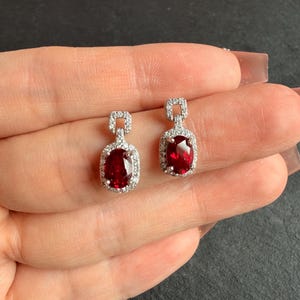 Puede incluir: Un par de pendientes colgantes con gemas de color rojo intenso de corte ovalado rodeadas por un halo de pequeñas piedras claras. Cada pendiente tiene un engaste rectangular incrustado con piedras claras en la parte superior.