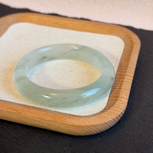 May include: A translucent jade bangle bracelet with a pale green hue. The smooth, polished surface of the bracelet reflects light, showcasing subtle variations in color. The jade bracelet is displayed on a wooden tray with a white linen insert.