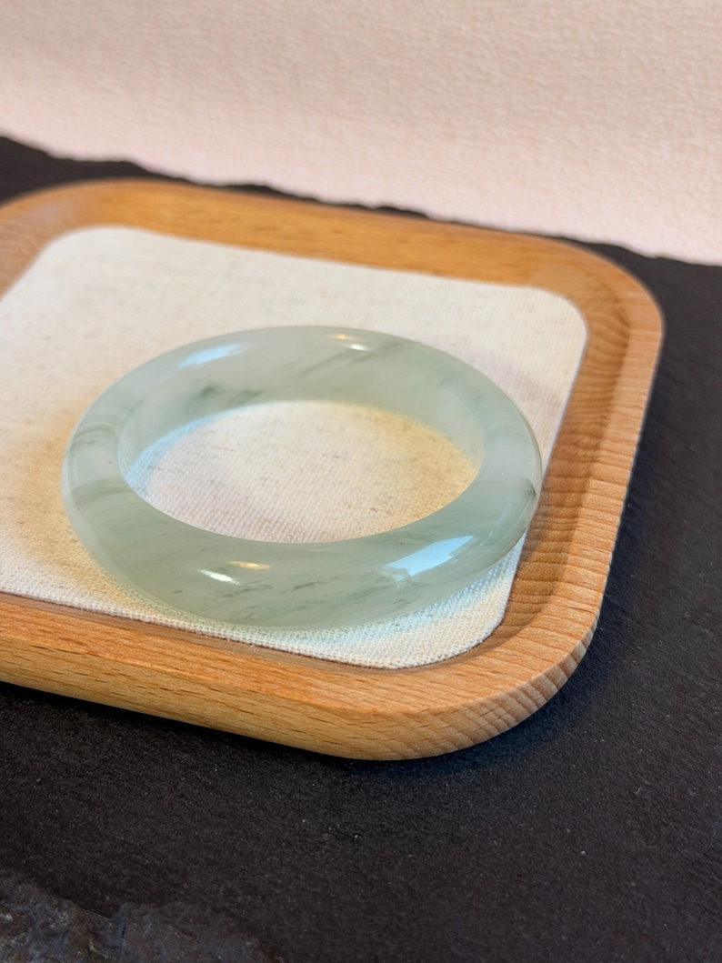 K&ouml;nnte beinhalten: Ein durchscheinendes Jade-Armreif-Armband in blassgr&uuml;ner Farbe. Die glatte, polierte Oberfl&auml;che des Armbands reflektiert das Licht und zeigt subtile Farbvariationen. Das Jade-Armband wird auf einem Holztablett mit wei&szlig;em Leinen-Einsatz pr&auml;sentiert.