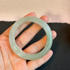 K&ouml;nnte beinhalten: Ein hellgr&uuml;nes Jade-Armreif-Armband. Das kreisf&ouml;rmige Armband wird in einer Hand gehalten, wodurch seine durchscheinende Qualit&auml;t hervorgehoben wird. Der Jade hat subtile Farbvariationen und eine polierte Oberfl&auml;che. Der Hintergrund ist neutral.