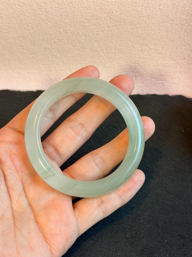 K&ouml;nnte beinhalten: Ein hellgr&uuml;nes Jade-Armreif-Armband. Das kreisf&ouml;rmige Armband wird in einer Hand gehalten, wodurch seine durchscheinende Qualit&auml;t hervorgehoben wird. Der Jade hat subtile Farbvariationen und eine polierte Oberfl&auml;che. Der Hintergrund ist neutral.