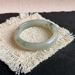 May include: A smooth, polished bangle bracelet crafted from translucent pale green stone with subtle darker green veining throughout. The circular jewelry piece features a rounded profile and a glossy finish, showcasing natural color variations.