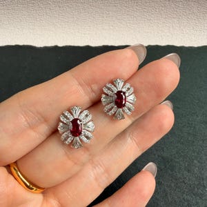 May include: A pair of silver-toned stud earrings featuring a central oval-cut deep red gemstone surrounded by a sunburst arrangement of small, clear, sparkling stones. The design creates a radiant, multi-faceted appearance with metallic accents.