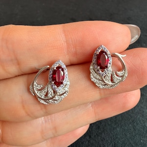 Pode incluir: Um par de brincos prateados com uma pedra preciosa central de corte oval em vermelho profundo. Cada brinco é rodeado por um halo de pequenas pedras violeta claro e uma estrutura curva em forma de folha incrustada com minúsculos cristais brancos.