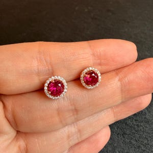 May include: A pair of stud earrings featuring a vibrant red gemstone surrounded by a halo of small, clear stones. The earrings have a gold-colored setting. The jewelry is displayed on a neutral background.