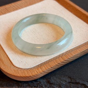 K&ouml;nnte beinhalten: Ein durchscheinendes Jade-Armreif-Armband in blassgr&uuml;ner Farbe. Die polierte Oberfl&auml;che reflektiert das Licht und zeigt die nat&uuml;rlichen Variationen des Steins. Das Armband wird auf einem Holztablett mit neutralem Stofffutter vor rosa Hintergrund pr&auml;sentiert.
