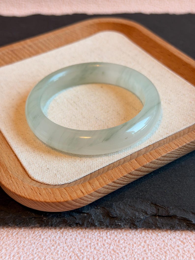 K&ouml;nnte beinhalten: Ein durchscheinendes Jade-Armreif-Armband in blassgr&uuml;ner Farbe. Die polierte Oberfl&auml;che reflektiert das Licht und zeigt die nat&uuml;rlichen Variationen des Steins. Das Armband wird auf einem Holztablett mit neutralem Stofffutter vor rosa Hintergrund pr&auml;sentiert.