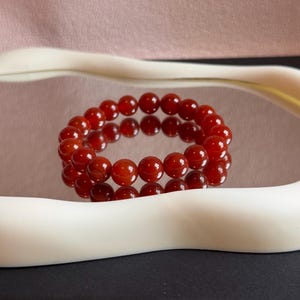 May include: A red carnelian beaded bracelet. The bracelet features polished, round beads. It's presented on a reflective surface, framed by white and black, against a pink backdrop.