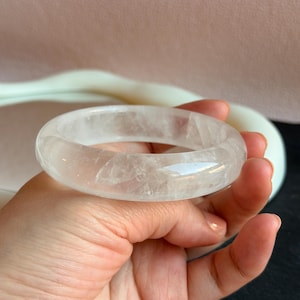 May include: A clear quartz bangle. The bracelet is a solid, rounded band with a translucent appearance. The quartz has subtle internal veining and a smooth, polished surface. The bracelet is held in a hand.