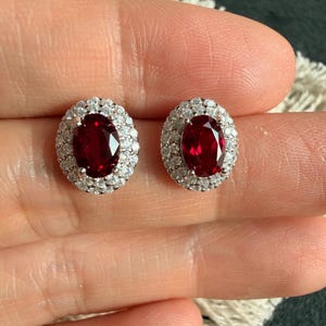 Puede incluir: Un par de pendientes de botón con piedras centrales de color rojo intenso de corte ovalado, rodeadas por un doble halo de pequeñas piedras blancas claras y brillantes engastadas en un marco de metal plateado pulido de aspecto elegante.