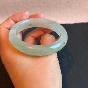 K&ouml;nnte beinhalten: Ein durchscheinendes, hellgr&uuml;nes Jade-Armreif-Armband. Das glatte, kreisf&ouml;rmige Armband weist subtile Farb- und Texturvariationen auf. Der Jade wird in einer Hand vor einem neutralen Hintergrund gehalten. Dieses elegante St&uuml;ck ist ein klassisches Schmuckst&uuml;ck.