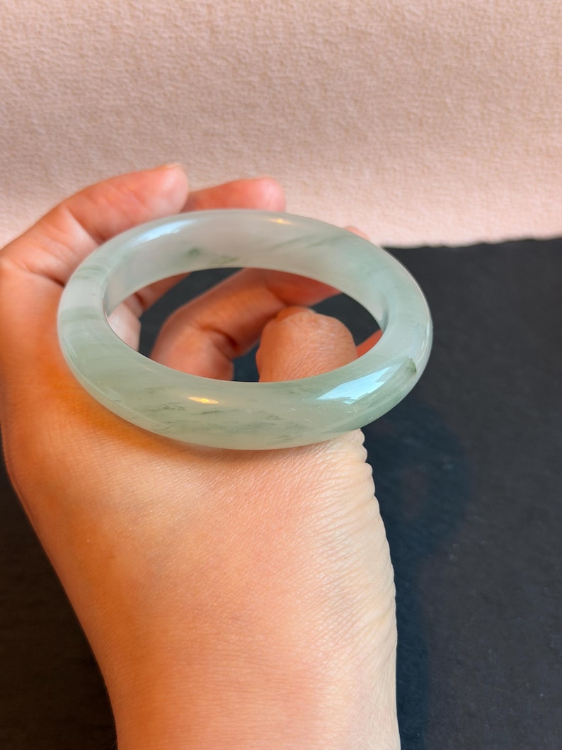 K&ouml;nnte beinhalten: Ein durchscheinendes, hellgr&uuml;nes Jade-Armreif-Armband. Das glatte, kreisf&ouml;rmige Armband weist subtile Farb- und Texturvariationen auf. Der Jade wird in einer Hand vor einem neutralen Hintergrund gehalten. Dieses elegante St&uuml;ck ist ein klassisches Schmuckst&uuml;ck.