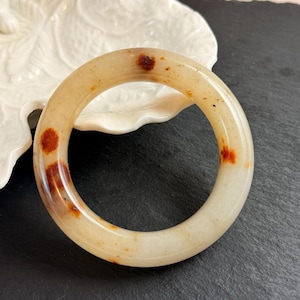 May include: A smooth, circular bangle bracelet crafted from translucent off-white stone, featuring distinct, irregular reddish-brown mineral inclusions scattered across its polished surface, creating a natural, mottled appearance.