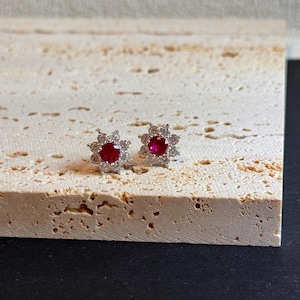 以下が含まれることがあります： 中央に赤い宝石、周囲に小さな透明な石をあしらったスタッドピアス。 ピアスは花柄で、明るい質感の表面に置かれています。 ジュエリーアイテムです。