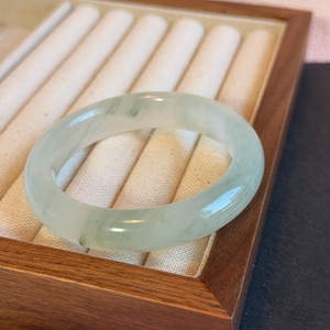 K&ouml;nnte beinhalten: Ein durchscheinendes, hellgr&uuml;nes Jadeit-Armreif-Armband. Die glatte, polierte Oberfl&auml;che reflektiert das Licht und zeigt die nat&uuml;rlichen Farbvariationen. Das Armband wird in einer Holzschmuckschatulle mit cremefarbenem Innenraum pr&auml;sentiert.