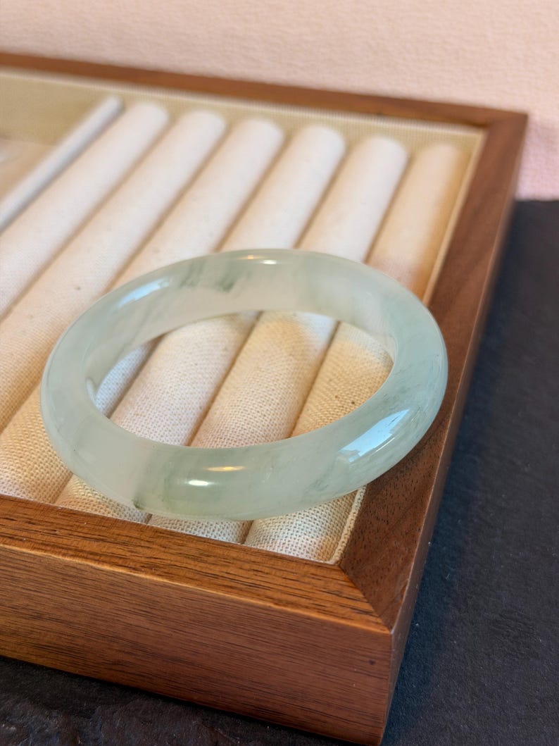 K&ouml;nnte beinhalten: Ein durchscheinendes, hellgr&uuml;nes Jadeit-Armreif-Armband. Die glatte, polierte Oberfl&auml;che reflektiert das Licht und zeigt die nat&uuml;rlichen Farbvariationen. Das Armband wird in einer Holzschmuckschatulle mit cremefarbenem Innenraum pr&auml;sentiert.