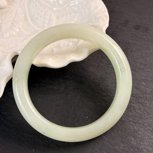 May include: A smooth, circular bangle bracelet crafted from pale, translucent light green stone. The bangle features a polished, glossy finish with subtle variations in color throughout the material, showcasing a simple, continuous band design.
