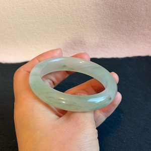 K&ouml;nnte beinhalten: Ein hellgr&uuml;nes Jade-Armreif-Armband. Die polierte Oberfl&auml;che des Armbands reflektiert das Licht und hebt seine nat&uuml;rlichen Variationen hervor. Der Jade ist hellgr&uuml;n mit dezenten, dunkleren gr&uuml;nen Adern. Kreisf&ouml;rmig.