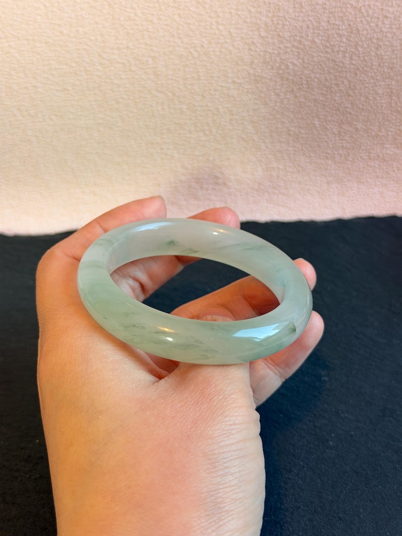 K&ouml;nnte beinhalten: Ein hellgr&uuml;nes Jade-Armreif-Armband. Die polierte Oberfl&auml;che des Armbands reflektiert das Licht und hebt seine nat&uuml;rlichen Variationen hervor. Der Jade ist hellgr&uuml;n mit dezenten, dunkleren gr&uuml;nen Adern. Kreisf&ouml;rmig.