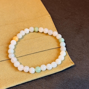 May include: White beaded bracelet with accents of orange and green beads. The bracelet is made of round, polished beads strung together on an elastic band. The bracelet is resting on a tan surface.