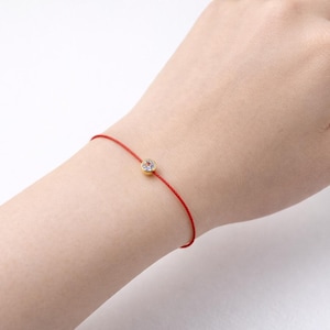May include: A delicate red string bracelet featuring a small, clear gemstone in a gold setting. The bracelet is worn on a wrist. The background is white.