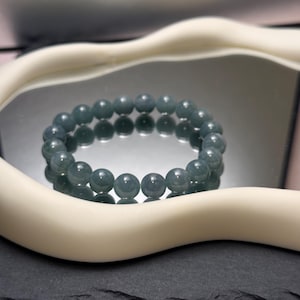 Może przedstawiać: Bransoletka z koralików składająca się z okrągłych, szaro-zielonych kamieni. Bransoletka jest prezentowana na odblaskowej powierzchni, obramowanej białym, zakrzywionym przedmiotem. Tło jest w kolorze jasnoróżowym.