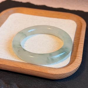 Op de afbeelding: Een doorschijnende jade armband met subtiele groene adering. De armband wordt gepresenteerd op een houten dienblad met een witte stoffen inzet, tegen een donkere achtergrond. De jade heeft een gladde, gepolijste afwerking.