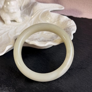 Puede incluir: Un brazalete de jade verde pálido. El brazalete circular se exhibe sobre un plato blanco en forma de hoja con una figura de conejo de cerámica. El fondo es una superficie oscura con un fondo rosa.