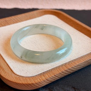 K&ouml;nnte beinhalten: Ein Armreif aus transluzentem Jade in blassgr&uuml;ner Farbe. Die polierte Oberfl&auml;che reflektiert das Licht und zeigt die nat&uuml;rlichen Variationen des Steins. Der Armreif liegt auf einem Holztablett mit einem neutralen Stofffutter. Durchmesser: ca. 6,5 cm.