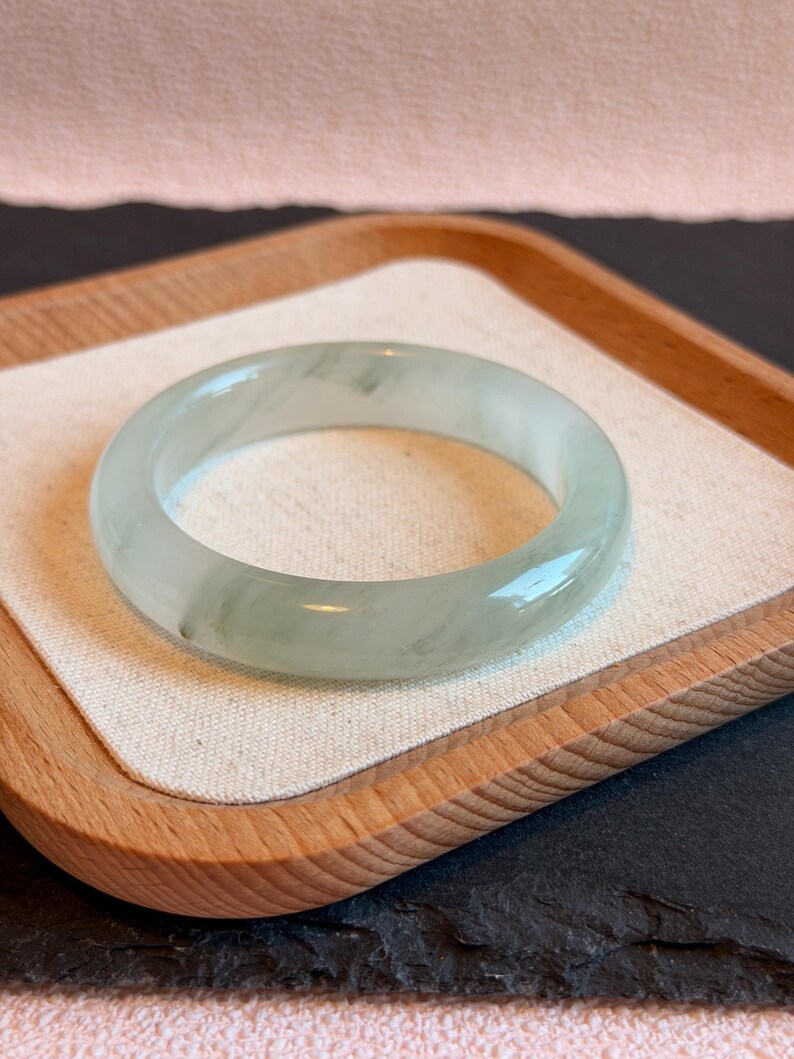 K&ouml;nnte beinhalten: Ein Armreif aus transluzentem Jade in blassgr&uuml;ner Farbe. Die polierte Oberfl&auml;che reflektiert das Licht und zeigt die nat&uuml;rlichen Variationen des Steins. Der Armreif liegt auf einem Holztablett mit einem neutralen Stofffutter. Durchmesser: ca. 6,5 cm.