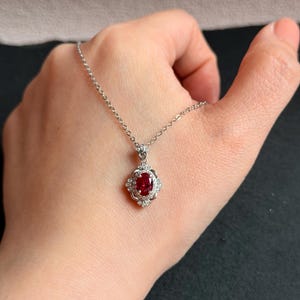 May include: A silver necklace featuring a pendant with a vibrant red gemstone at its center. The gemstone is surrounded by a halo of smaller, clear stones. The pendant hangs from a delicate silver chain.
