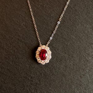 May include: A silver necklace with a pendant featuring a large oval ruby surrounded by a halo of smaller, clear gemstones. The ruby is a deep red colour, and the necklace is set against a dark background.