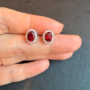 Peut inclure: Une paire de boucles d'oreilles à clous en rubis. Chaque boucle d'oreille présente une pierre précieuse rouge vif de forme ovale, entourée de petits cristaux transparents. Les boucles d'oreilles sont serties dans un métal argenté.
