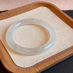 Könnte beinhalten: Ein hellgrünes Jade-Armreif-Armband. Das kreisförmige Armband wird auf einem Holztablett mit einem beigefarbenen Leinen-Einsatz präsentiert. Der Jade weist subtile Farb- und Texturvariationen auf und reflektiert das Licht. Durchmesser: ca. 6,5 cm.