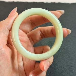May include: A smooth, circular bangle bracelet crafted from a solid piece of pale, translucent light green stone. The bangle features a polished, rounded surface with a consistent thickness throughout its entire circumference, showcasing a soft, uniform color.