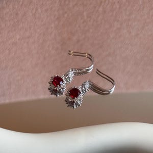 Könnte beinhalten: Ein Paar silberne Ohrringe mit einem roten Edelstein in der Mitte, umgeben von kleineren, klaren Steinen. Die Ohrringe haben ein florales Design und einen gebogenen Haken.
