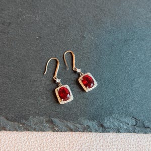 May include: Dangle earrings with a vivid red gemstone in a rectangular setting of small, sparkling white stones. The earrings have a hook closure. The earrings are displayed on a dark grey surface.