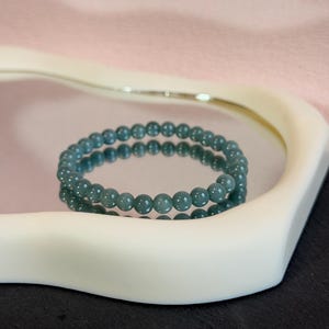 Puede incluir: Una pulsera de cuentas de color verde azulado descansa sobre una bandeja blanca con una superficie de espejo, que refleja la pulsera. La pulsera está compuesta por pequeñas cuentas redondas. El fondo presenta una superficie rosa y negra.