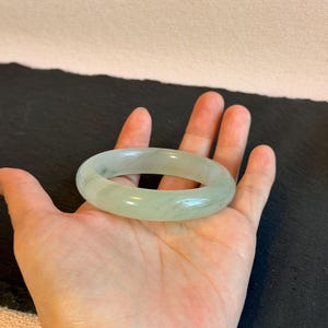 K&ouml;nnte beinhalten: Ein hellgr&uuml;nes Jade-Armreif-Armband. Das glatte, polierte Armband ist oval geformt und liegt auf einer offenen Handfl&auml;che. Der Jade hat eine durchscheinende Qualit&auml;t mit subtilen Farbvariationen. Der Hintergrund ist eine dunkle Oberfl&auml;che.