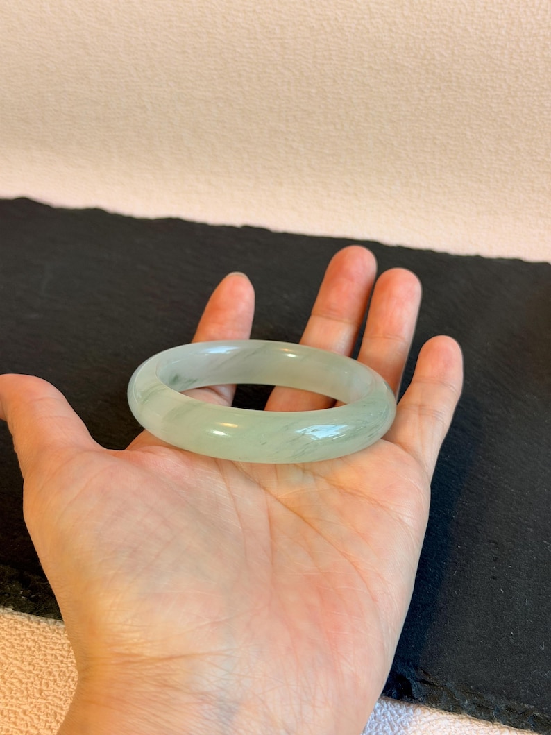 K&ouml;nnte beinhalten: Ein hellgr&uuml;nes Jade-Armreif-Armband. Das glatte, polierte Armband ist oval geformt und liegt auf einer offenen Handfl&auml;che. Der Jade hat eine durchscheinende Qualit&auml;t mit subtilen Farbvariationen. Der Hintergrund ist eine dunkle Oberfl&auml;che.