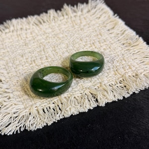 Puede incluir: Dos anillos de color verde oscuro, lisos y pulidos, con un perfil de banda grueso y redondeado. Los anillos presentan un acabado brillante y un color verde bosque profundo y sólido con sutiles variaciones naturales en la textura.