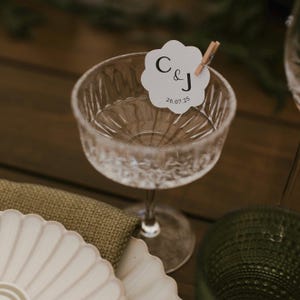 May include: A clear, etched glass champagne coupe with a white floral-shaped tag clipped to the rim. The tag has the letters "C & J" and the date "26.07.25" printed on it. The coupe sits on a wooden surface.