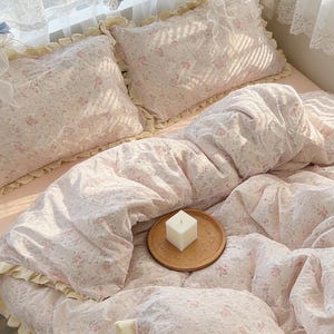 May include: A bed with a floral patterned comforter and pillow set in shades of pink and white. The bedding has a ruffled edge. A small wooden tray with a white candle sits on the comforter. The text "WhatiFinds" is visible in the bottom left corner.